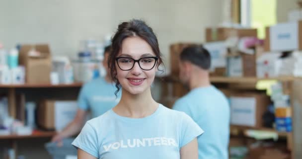 穿着蓝色志愿衬衫和眼镜的年轻貌美女子在慈善基金会仓库中摆姿势的画像 研究模糊背景的同事小组 — 图库视频影像