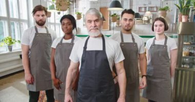 Portrait of mixed-race different male and female workers in aprons standing in cozy cafeteria looking at camera in good mood Restaurant business industry Senior cafe owner with young employees at work
