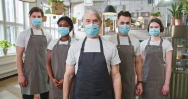 Portrait of mixed-race male and female workers in aprons and medical masks standing in cozy restaurant looking at camera in good mood waiting for customers. Business industry. Work in quarantine