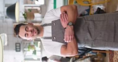Vertical video of cheerful young Caucasian man worker in apron standing in restaurant, looking at camera and smiling at work. Male barista in coffee shop with smile on face. Small business owner