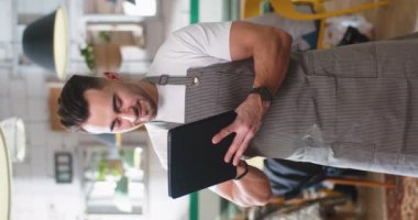 Portrait of young handsome Caucasian joyful man worker in apron typing on tablet standing in coffee house and smiling. Vertical orientation of happy cafe owner browsing on device. Restaurant service