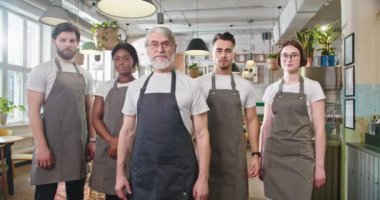 Camera approaching joyful multi-ethnic colleagues in aprons in restaurant, looking at camera and smiling. Portrait of senior and young diverse workers in cafeteria with smile on faces, work concept