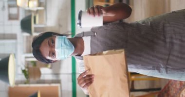 Afro-Amerikalı genç güzel bir kadının portresi. Tıp maskesinde duruyor. Kahve paketine bakıyor. Paket siparişle birlikte.