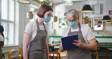 Portrait of Caucasian senior male restaurant owner in medical mask and apron speaking with young beautiful female worker in cafeteria at work writing down something, covid-19, job concept