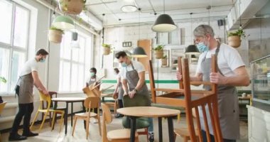 Mixed-race male and female workers working in cafeteria in medical masks cleaning in restaurant and putting chairs on tables. People work in coffee house in quarantine, no customers, covid-19