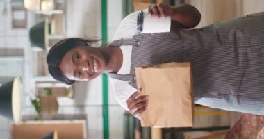 Portrait of happy beautiful young African American woman barista smiling while standing in restaurant looking at camera holding take away order and coffee-to-go in hands. Vertical video small business