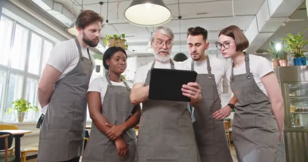 多人種の若い男性と女性従業員の作業計画と議論画面を見てタブレットをタップエプロンで忙しいシニア白人男性レストランのオーナーの肖像画 事業概念 — ストック動画
