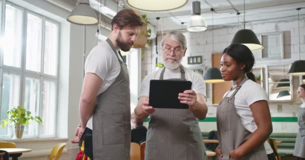 若い多民族のウェイターに何かを説明する同僚と話すデバイスアプリを使用してタブレット上でタップコーヒーハウスに立っている古い素敵な白髪の男カフェマネージャー 食品産業事業 — ストック動画