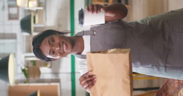 アフリカ系アメリカ人の美しい若い女性コーヒーショップのオーナーの肖像画 笑顔で紙袋とコーヒーの注文を取り除き 屋内でカメラを見ながら行きます 垂直方向のビデオ — ストック動画