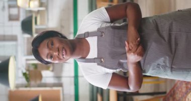 Afro-Amerikan genç, güzel, mutlu kadın işçinin portresi. Önlüklü, kahve dükkanında duruyor ve gülümsüyor, kameraya bakıyor ve konuşuyor. Video blogu kaydediliyor. Dikey yönelim videosu
