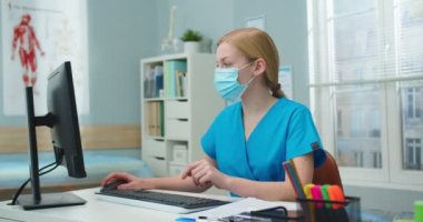 Hastanede bilgisayarla çalışan kendine güvenen genç bir doktor. Beyaz kadın hemşire koruyucu maske takıyor ve ofisteki kameraya bakıyor. Coronavirus, tıp konsepti.