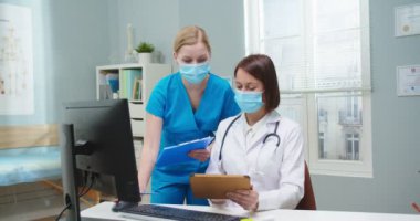 Koruyucu maskeli profesyonel kadın doktor ve rapor veren genç asistan. Kafkasyalı yetişkin bir kadın tablet tutuyor ve mavi üniformalı hemşireye tıbbi verileri dikte ediyor. Tıp konsepti.