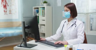Hastane ofisinde bilgisayarla çalışan beyaz bir kadının portresi. Tıbbi maske takmış yetişkin bir kadın doktor kameraya bakıyor, daktilo ediyor. Tıp, teknoloji konsepti.