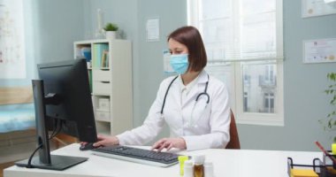 Koruyucu maskeli yetişkin kadın doktor hastane ofisinde bilgisayar üzerinde çalışıyor. Mavi üniformalı genç bir hemşire odaya giriyor ve patrona sağlık raporlarını kontrol etmesini söylüyor. İlaç, salgın, sağlık.