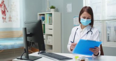 Tıbbi koruyucu maskeli çekici kadın doktor hastane ofisinde belgelerle çalışıyor. Beyaz cüppeli yetişkin bir kadın dosya kağıtlarına veri dolduruyor ve bilgisayar kullanıyor. Tıp konsepti.