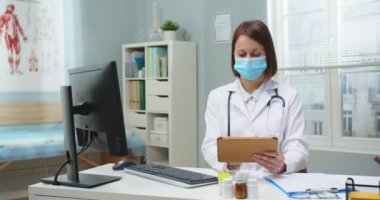 Masada oturup tablet kullanan, tıbbi koruyucu maskeli beyaz bir kadın. Yetişkin kadın doktor görüntüyü kaydırıyor, hastanedeki cihazdan e-posta yolluyor ve kontrol ediyor. Teknoloji, tıp.