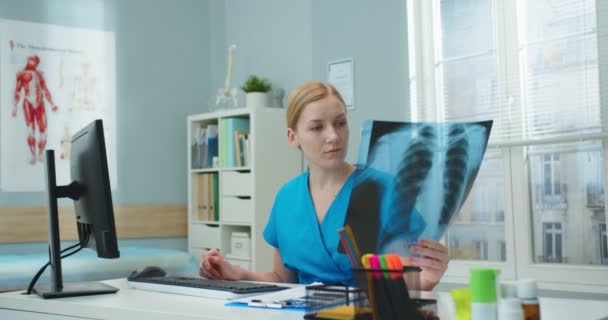 年轻漂亮的希望工作者坐在桌旁看着X光扫描 身穿蓝色医疗服的专业白人女护士记录了有关疾病的报告 — 图库视频影像