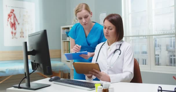 病院のオフィスでアシスタンと文書化する女性の医師を魅了 白いガウンの若い白人看護師がレポートを作成し 女性に指示を与える 職業コンセプト — ストック動画