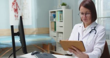 Hastanedeki nöbetçi odasında dijital tablet üzerinde çalışan kendine güvenen bir kadın doktor. Gözlüklü yetişkin bir beyaz kadın cihazla ilgili tıbbi raporlar hazırlıyor ve bilgisayardan verileri kontrol ediyor. Teknoloji konsepti.