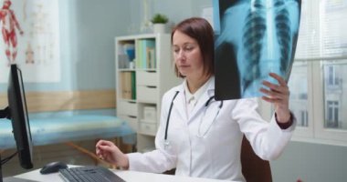 Masada çalışan profesyonel kadın doktor, röntgene bakıyor ve verileri dolduruyor. Beyaz tıbbi önlüklü yetişkin bir kadın bilgisayarda tarama sonuçlarını belgeleyerek rapor veriyor. Teknoloji.