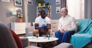 Beyaz saçlı, son sınıf öğrencisi, Afrikalı Amerikalı genç erkek bir doktor ya da hemşire ile ev ziyaretinde konuşuyor. Sağlık sorununu açıklıyor. Kanepede oturuyor ve doktor bilgi yazıyor.