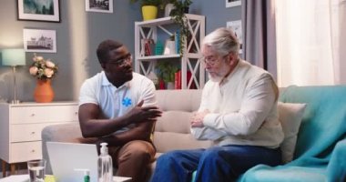 Ciddi bir şekilde ilgilenen Afrikalı Amerikalı erkek doktor. Evde tıbbi konsültasyonda, oturma odasındaki kanepede otururken dizüstü bilgisayarında yazı yazan Kafkasyalı yaşlı bir hastayla konuşuyor.