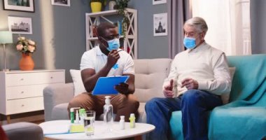Karantinaya ev ziyareti sırasında panoya yazı yazan Kafkasyalı yaşlı bir hastayla konuşan tıbbi maskeli Afrikalı Amerikalı genç erkek doktor, sağlık kontrolü konsepti