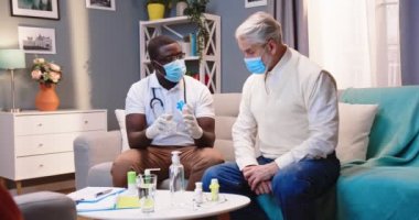Sağlık çalışanı Afrikalı Amerikalı genç doktor, karantinada tıbbi maske takarak ve sağlık kontrolü yaparak beyaz saçlı yaşlı bir hastayı evinde ziyaret ediyor.