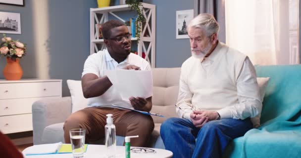 非洲裔美国青年专业全科医生在家里拜访白种人老年男性病人 谈论治疗结果和坐在客厅沙发上的治疗 医生探访 医疗服务 — 图库视频影像