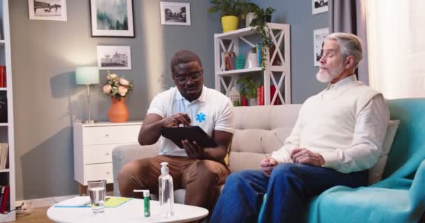 在家庭医疗会诊期间 年轻而集中的非洲裔美国男性专业医生与坐在沙发上的白种人老年男性患者交谈 在平板电脑上显示出打字结果 — 图库视频影像