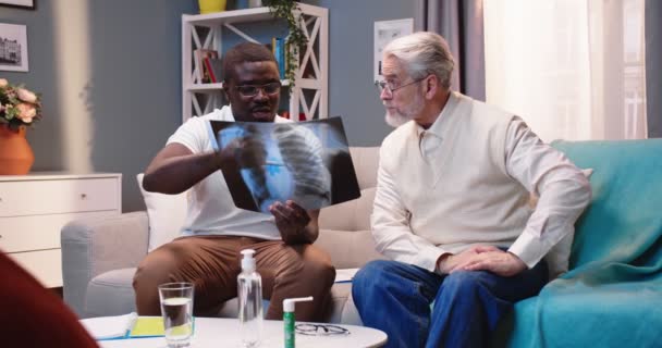 非洲裔美国年轻男性专业医生在家中拜访白种人老年男性患者 并在客厅沙发上展示X光扫描结果 保健访问 家庭治疗 — 图库视频影像