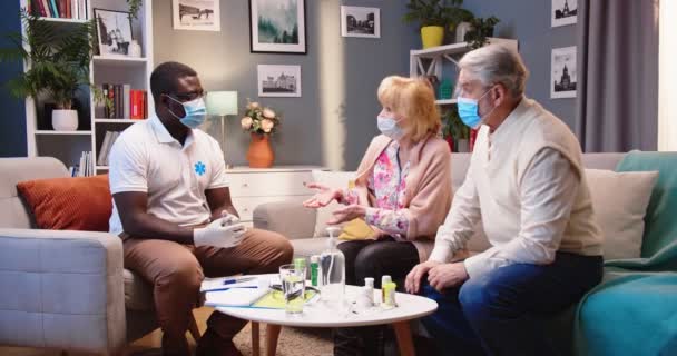 古い患者の女性と男性のカップルは リビングルームでソファーに座っている医療マスクの若いアフリカ系アメリカ人男性の専門医と話し 自宅で医療相談しました ホームケアのコンセプト — ストック動画
