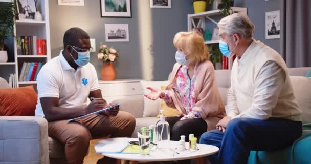 与白种人交谈的非裔美国青年医生或护理人员已婚夫妇 他们都戴着口罩 在家中看病时写下症状 保健概念 — 图库视频影像
