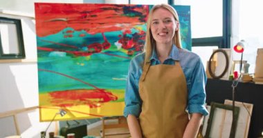 Portrait of cheerful young Caucasian beautiful talented woman artist in apron standing in studio workshop looking at camera and smiling in good mood, female painter with happy face