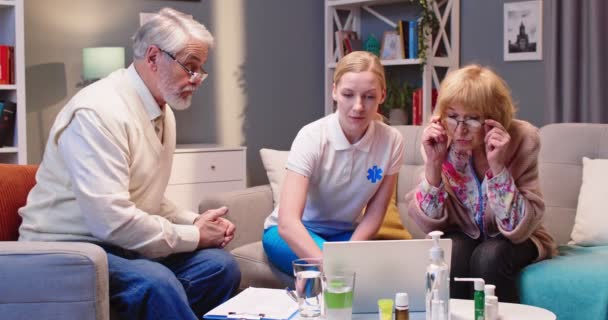 若い女性のプロの看護師は 古い退職した患者とソファに座って 家庭訪問中にラップトップコンピュータにタイピングする分析結果を説明し 示しています — ストック動画