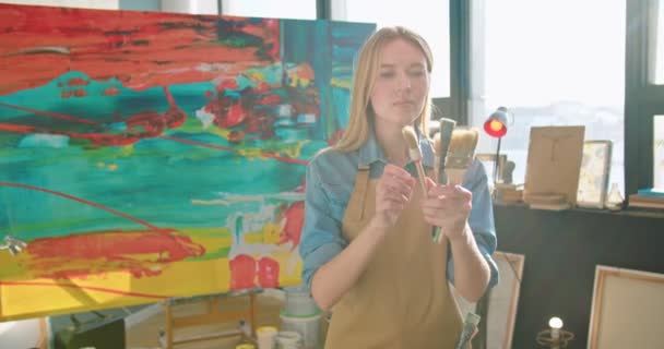 才华横溢的年轻白种人专业女艺术家站在艺术作坊围裙选择画笔 看着相机 在灿烂的阳光下微笑 画家的概念 — 图库视频影像
