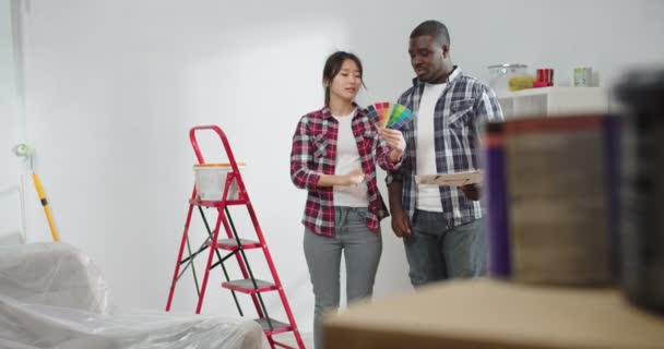 若い多民族カップル アフリカ系アメリカ人の夫とアジアの妻は 新しいアパートの改装中に色を選択する部屋に立って コンセプトを装飾し 再設計し 家を改装 — ストック動画