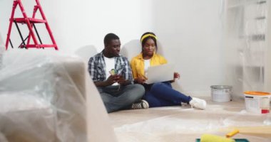 Afro-Amerikalı genç evli çift, dizüstü bilgisayarla mesajlaşarak oda tasarımı ve duvar rengini seçerken yeni bir dairede, yenileme ve onarım konseptinde oturuyorlar.