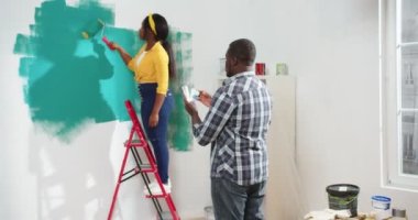 Yetişkin Afro-Amerikan bir erkek odada dikilip boya rengini seçerken genç ve güzel karısının duvarı nasıl yeşil renge boyadığını görüyor. Ev onarımı yapan aile çifti, ev yenileme konsepti.