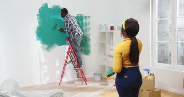 Afrika kökenli Amerikalı bir kadın odada durmuş yeşil renkli duvar boyayan kocaya bakıyor. Ev onarım konsepti sırasında kameraya gülümseyen genç bir eşin portresi.