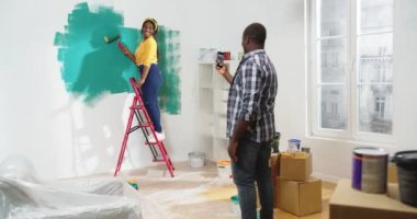 Akıllı telefon kamerasına poz veren neşeli genç bir kadın merdiven üzerinde durup duvarı yeşile boyuyor ve gülümsüyor. Afrikalı Amerikalı kocanın cep telefonuyla fotoğraf çekmesi, tamir konsepti