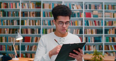 Gözlüklü Latin genç adam kütüphanede elinde belgelerle duruyor, not alıyor ve bir şeyler yazıyor. Bazı kağıtları kontrol ederken bibliyotheca 'da okuyan erkek öğrenci.