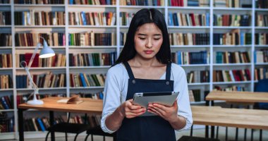 Kütüphanede tabletle çalışan ve gülümseyen zeki Asyalı Yang kadınının yakın plan portresi. Arkaplanda kitaplık.