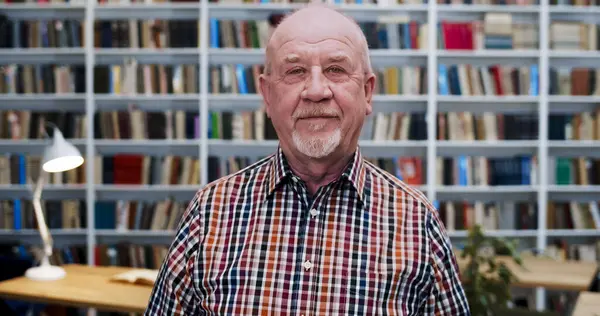 Portrait shot of old Caucasian bald man in motley shirt looking at camera with slight smile in library. Male professor or worker of bibliotheca smiling. Books shelves on background. Senior teacher.