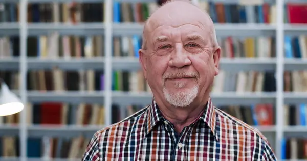 Portrait of old Caucasian bald man in motley shirt looking to camera and smiling in library. Close up of male professor or cheerful worker of bibliotheca. Books shelves behind. Senior smiled teacher.