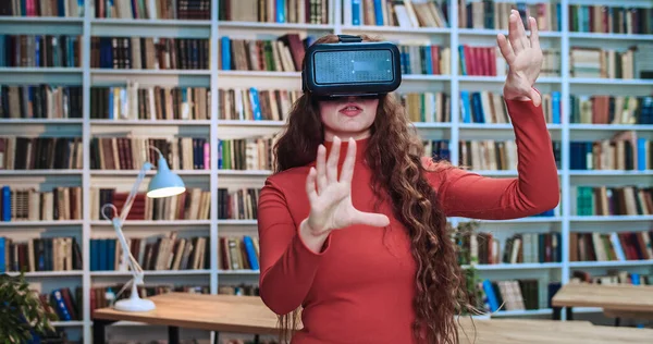Close-up portrait of beautiful young red head woman wearing virtual reality headset touch something using modern 3D vr glasses in library next to book rack.