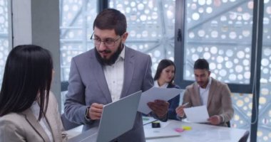 Gözlüklü beyaz adam elinde dizüstü bilgisayarla Asyalı genç bayan çalışana belgeler gösteriyor. Erkek ve bayan ofis çalışanları sohbet ediyor ve iş için çözüm arıyorlar..