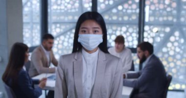 Kamera, tıbbi maskeli genç Asyalı kadın ofis çalışanına odaklanıyor ve kameraya bakıyor. Arka planda iş toplantısı. Salgın sırasında şirket çalışanlarının portresi.