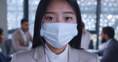 Ofiste maske takmış kameraya bakan Asyalı beyaz yakalı genç bayan ofis çalışanının yüzünü kapatın. Arka planda iş toplantısı. Kadın şirket çalışanının portresi. Koronavirüs salgını.