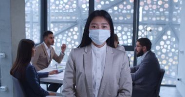 Ofiste duran ve kameraya bakan tıbbi maskeli genç Asyalı kadın ofis çalışanı portresi. Arka planda iş toplantısı. Coronavirus salgını sırasında çalışan kadın..